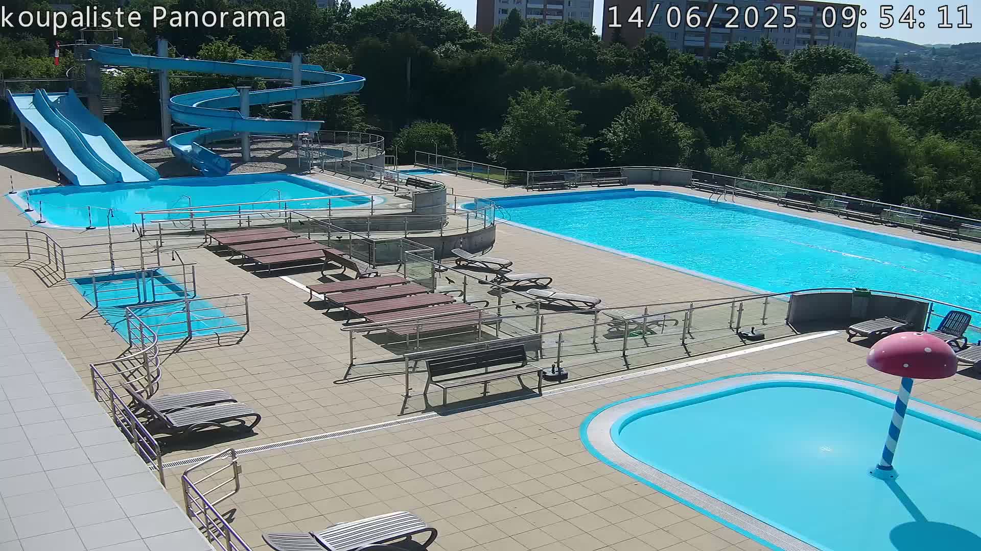 An outdoor pool complex with multiple pools, water slides, lounge chairs, and a small mushroom-shaped water feature on a sunny day.