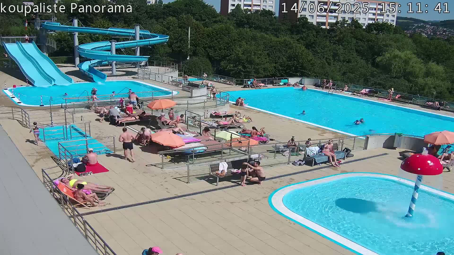 An outdoor pool complex on a sunny day features two large swimming pools, water slides, and numerous people relaxing and swimming.