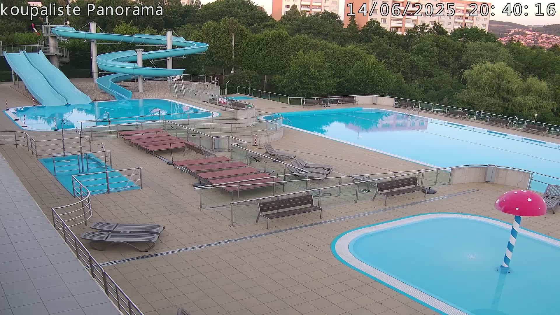An outdoor pool complex on a sunny day features two large swimming pools, water slides, and numerous people relaxing and swimming.
