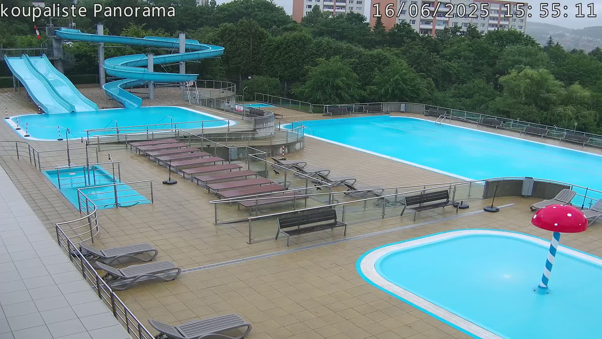 An outdoor pool complex with two large pools, a small wading pool, two water slides, and numerous lounge chairs under overcast skies.