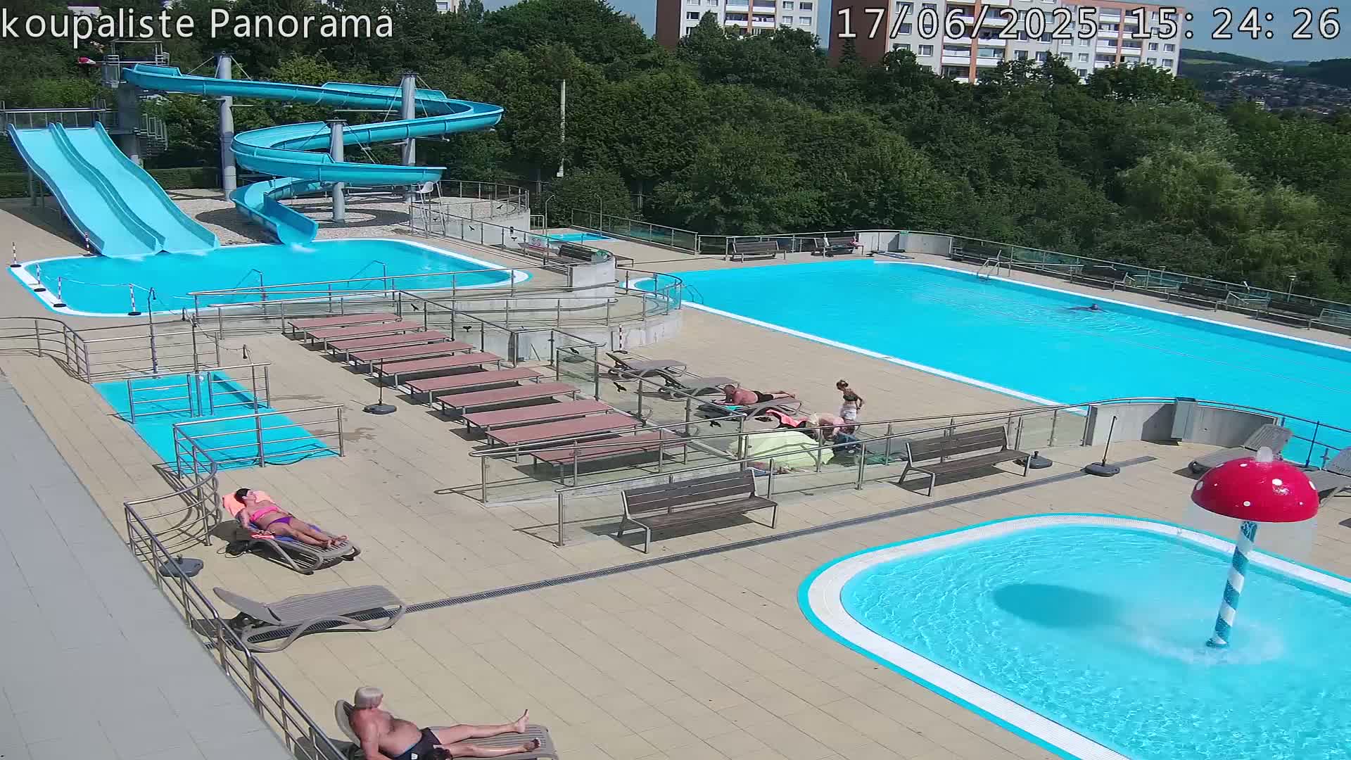 An outdoor pool complex on a sunny day features two large pools, several smaller pools, water slides, lounge chairs, and people relaxing.