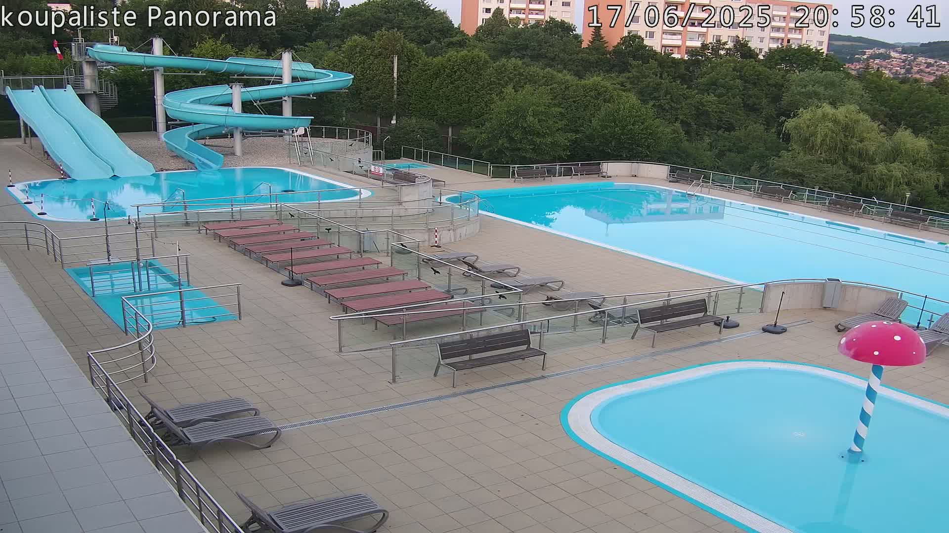 An outdoor swimming pool complex with two large pools, several smaller pools, water slides, lounge chairs, and benches under an overcast sky.