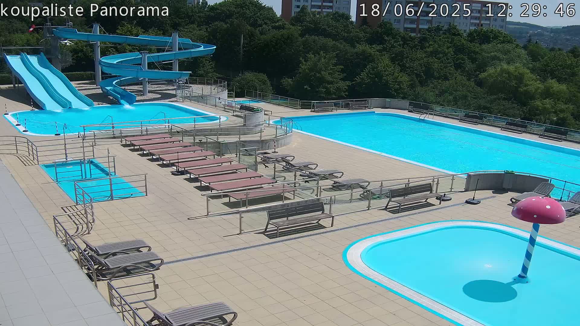 An outdoor pool complex on a sunny day features two large pools, several smaller pools, water slides, lounge chairs, and benches.