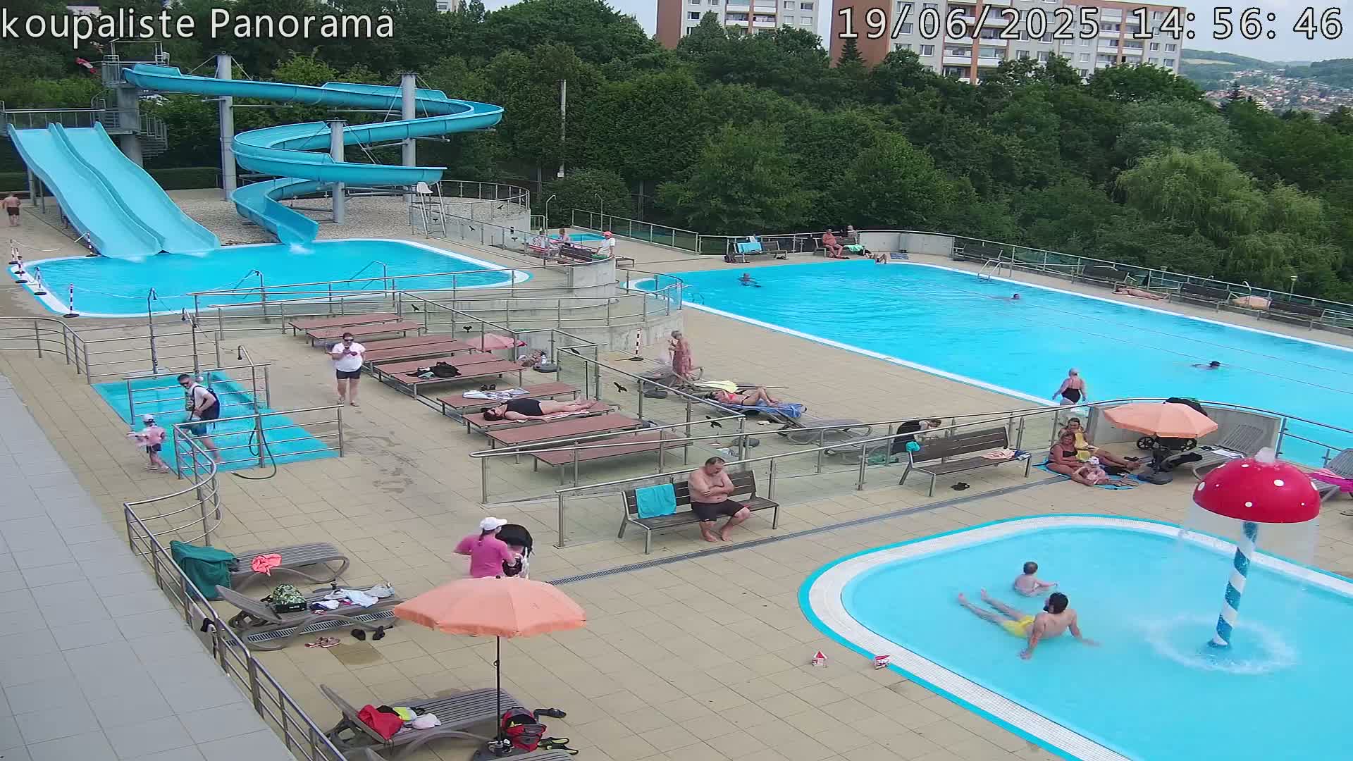 An outdoor pool complex with two water slides, a large swimming pool, a smaller children's pool with a mushroom-shaped fountain, and numerous people lounging on deck chairs.