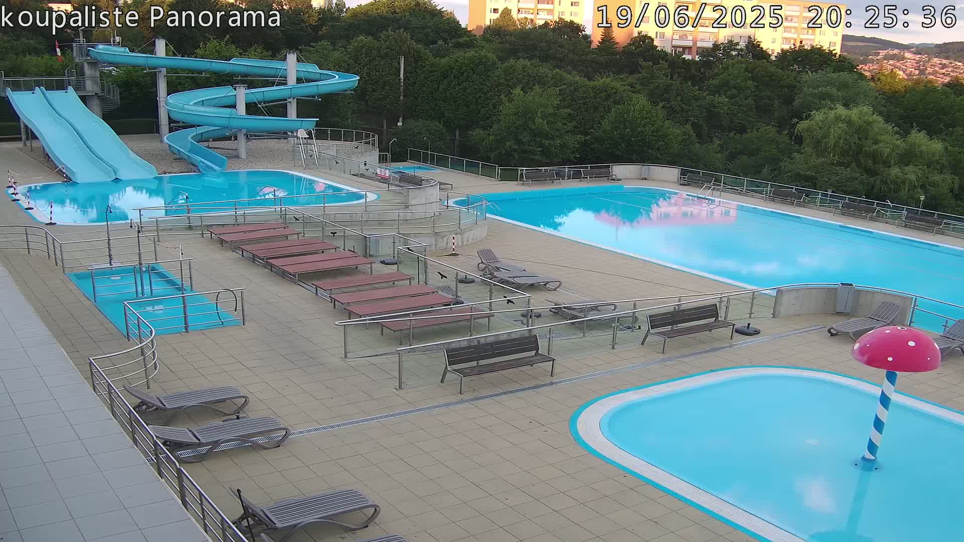 An outdoor swimming pool complex with two large pools, a smaller children's pool, two slides, and numerous lounge chairs under a twilight sky.