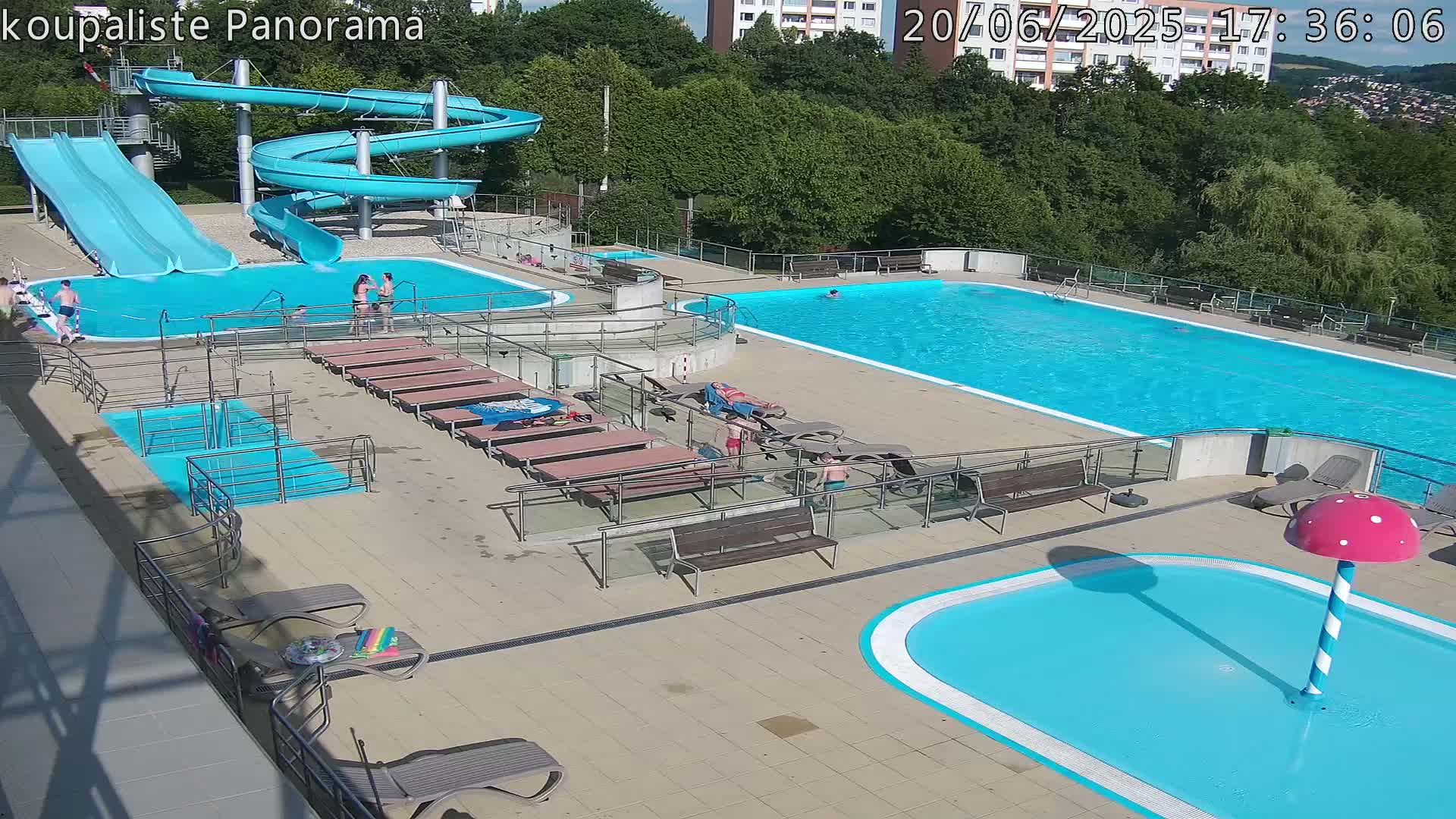 An outdoor swimming complex on a sunny day features a large pool, a smaller children's pool with a mushroom-shaped water feature, two slides, and several lounge chairs surrounding the pools.