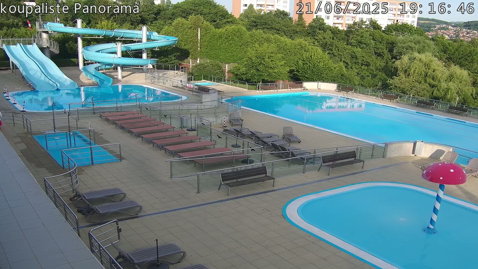 An outdoor pool complex with multiple pools, water slides, lounge chairs, and a mushroom-shaped water feature, under a mostly sunny sky.