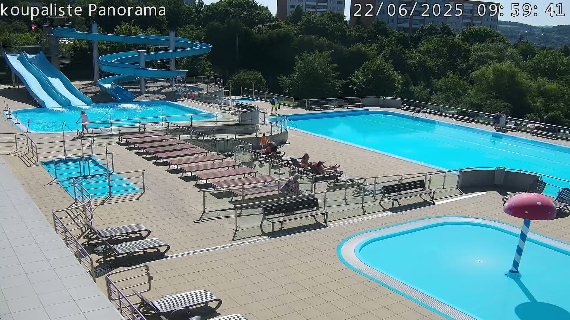 An outdoor pool complex on a sunny day features a large lap pool, a smaller pool with a mushroom-shaped water feature, several slides, and lounge chairs surrounding the pools.
