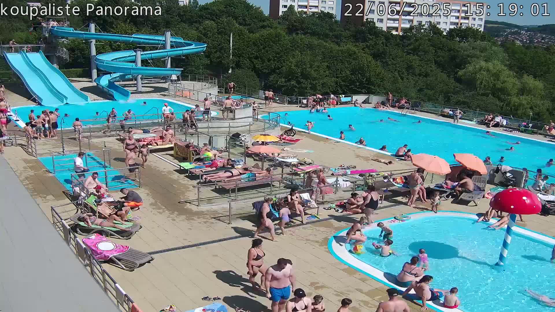 A sunny outdoor swimming pool complex with multiple pools, slides, and many people enjoying the sun and water.
