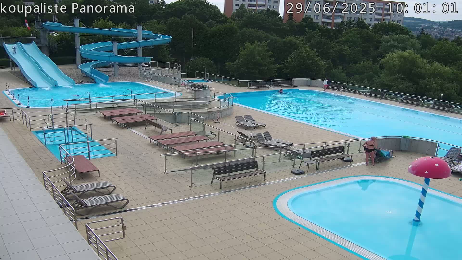 An outdoor pool complex with two large swimming pools, several smaller pools, water slides, lounge chairs, and benches, under a partly sunny sky.
