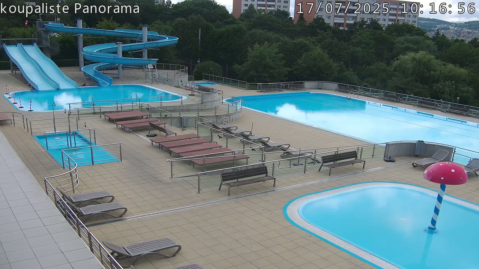 An outdoor pool complex with multiple pools, water slides, lounge chairs, and a small mushroom-shaped water feature on a sunny day.