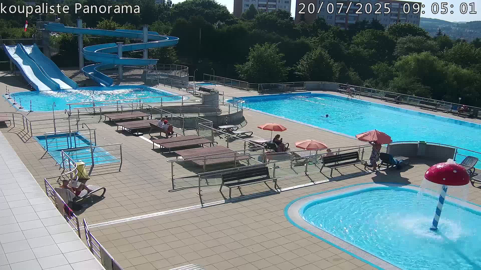 An outdoor pool complex on a sunny day features several pools of varying sizes, water slides, lounge chairs, and umbrellas, with people relaxing and swimming.