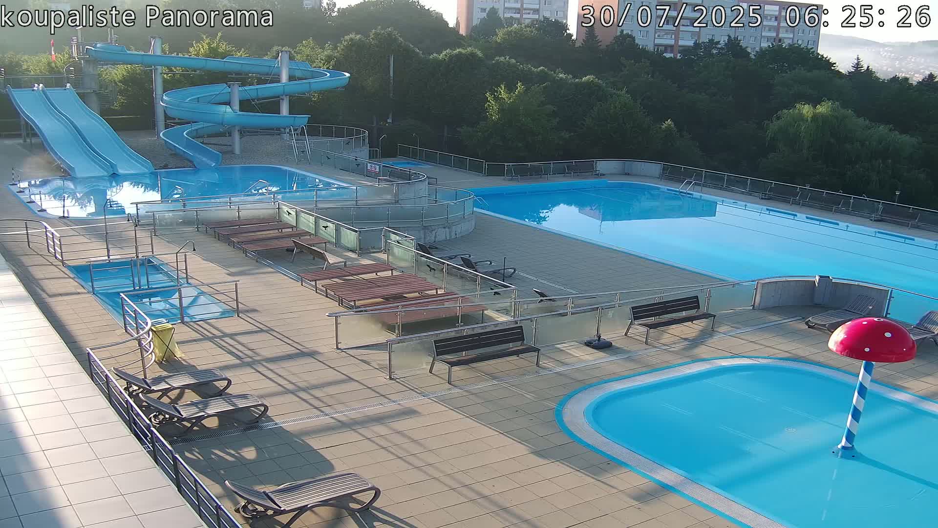 An outdoor pool complex on a sunny day features several pools of varying sizes, water slides, lounge chairs, and umbrellas, with people relaxing and swimming.