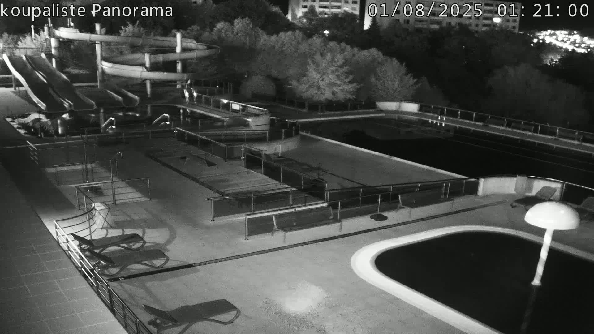 An empty outdoor pool area at night with several water slides, lounge chairs, and surrounding trees is illuminated by artificial light.