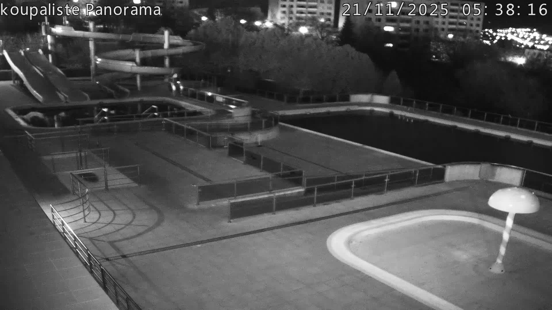 A vacant outdoor swimming pool complex featuring light blue water slides, several pools (some empty, one filled with murky green water), and a red mushroom water feature is seen under an overcast sky, with bare trees and apartment buildings in the background.
