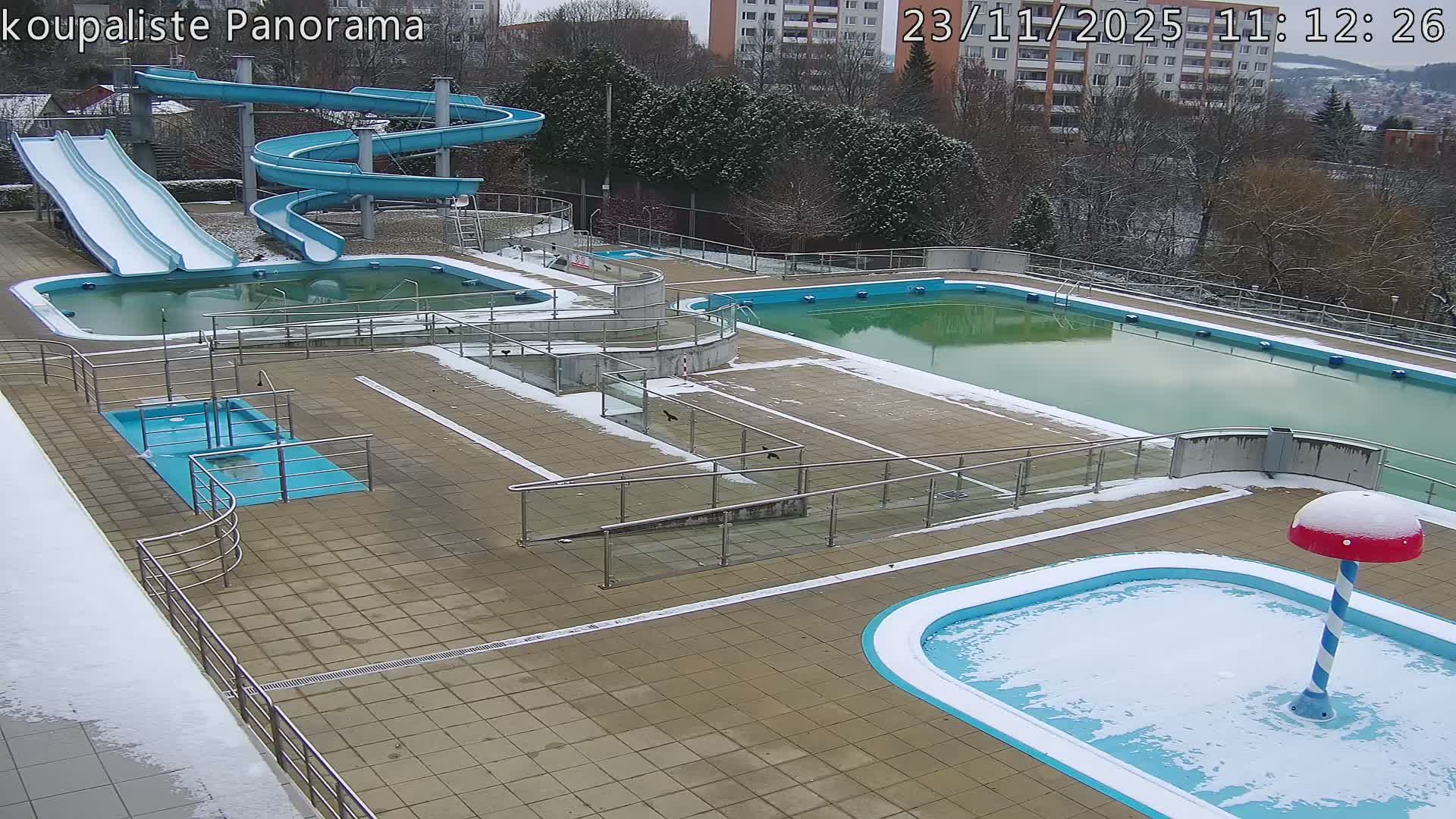 An outdoor swimming pool complex, featuring various slides and multiple pools with differing water and frozen surfaces, is partially covered in snow and ice, reflecting cold, snowy winter weather.