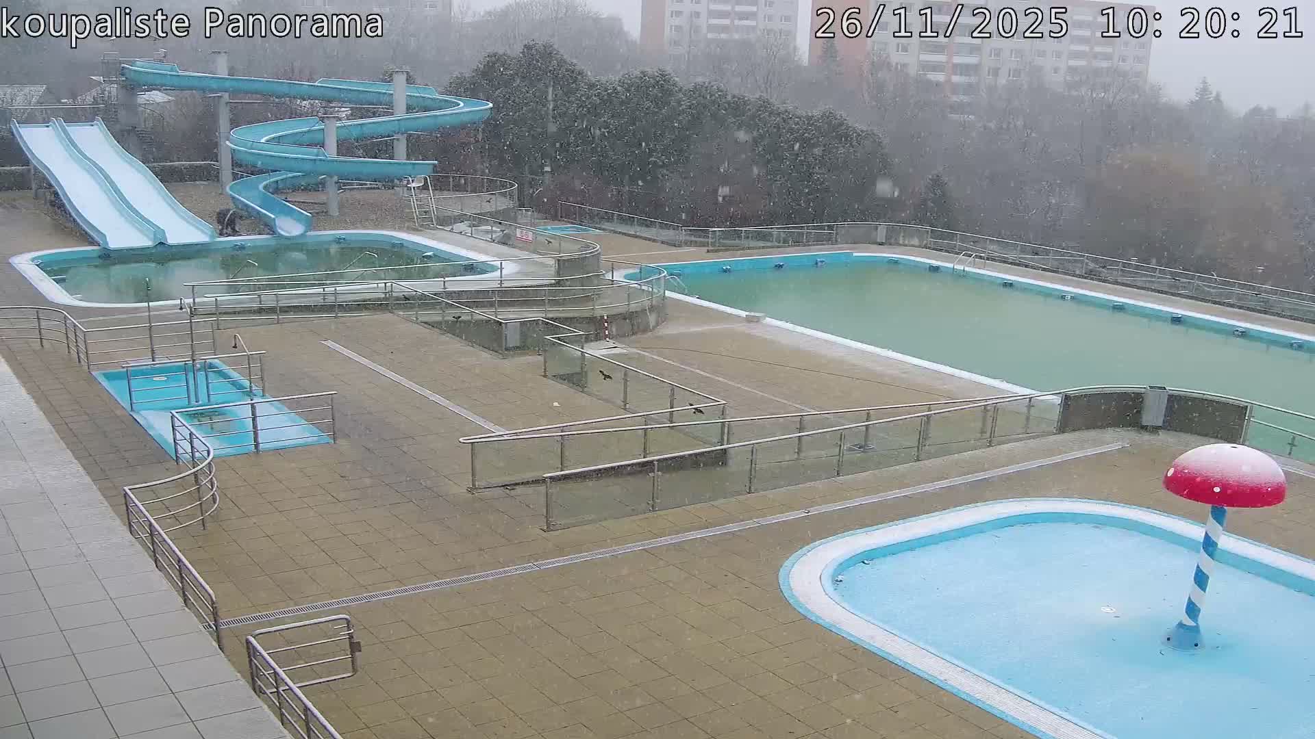 A deserted outdoor swimming pool complex, featuring various empty or murky pools and water slides, is seen under a steady snowfall.