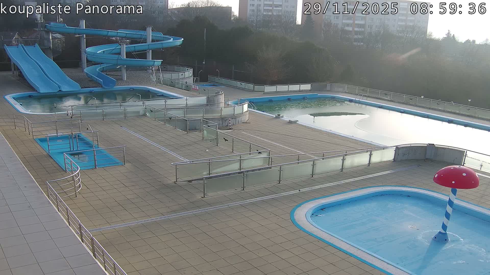 An outdoor swimming pool complex with various blue water slides, partially filled or drained pools, and a red mushroom-shaped water feature sits under a bright, possibly hazy morning sky.