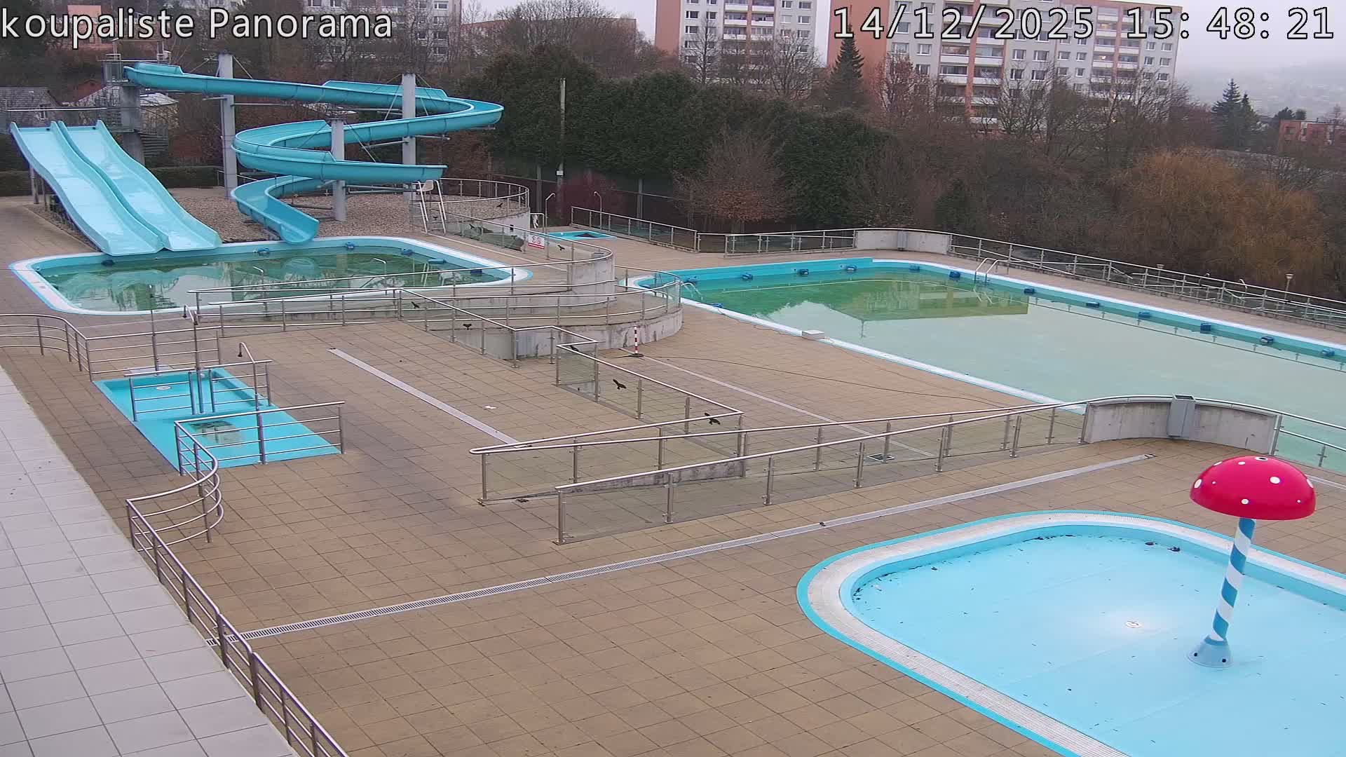 An empty outdoor swimming pool complex featuring multiple pools, some murky and some clearer, along with blue water slides and a mushroom water feature, is depicted under an overcast sky.