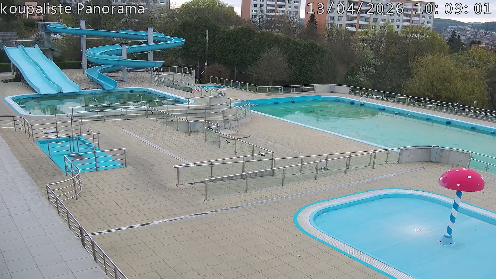 An empty outdoor swimming pool complex featuring multiple pools, some murky and some clearer, along with blue water slides and a mushroom water feature, is depicted under an overcast sky.