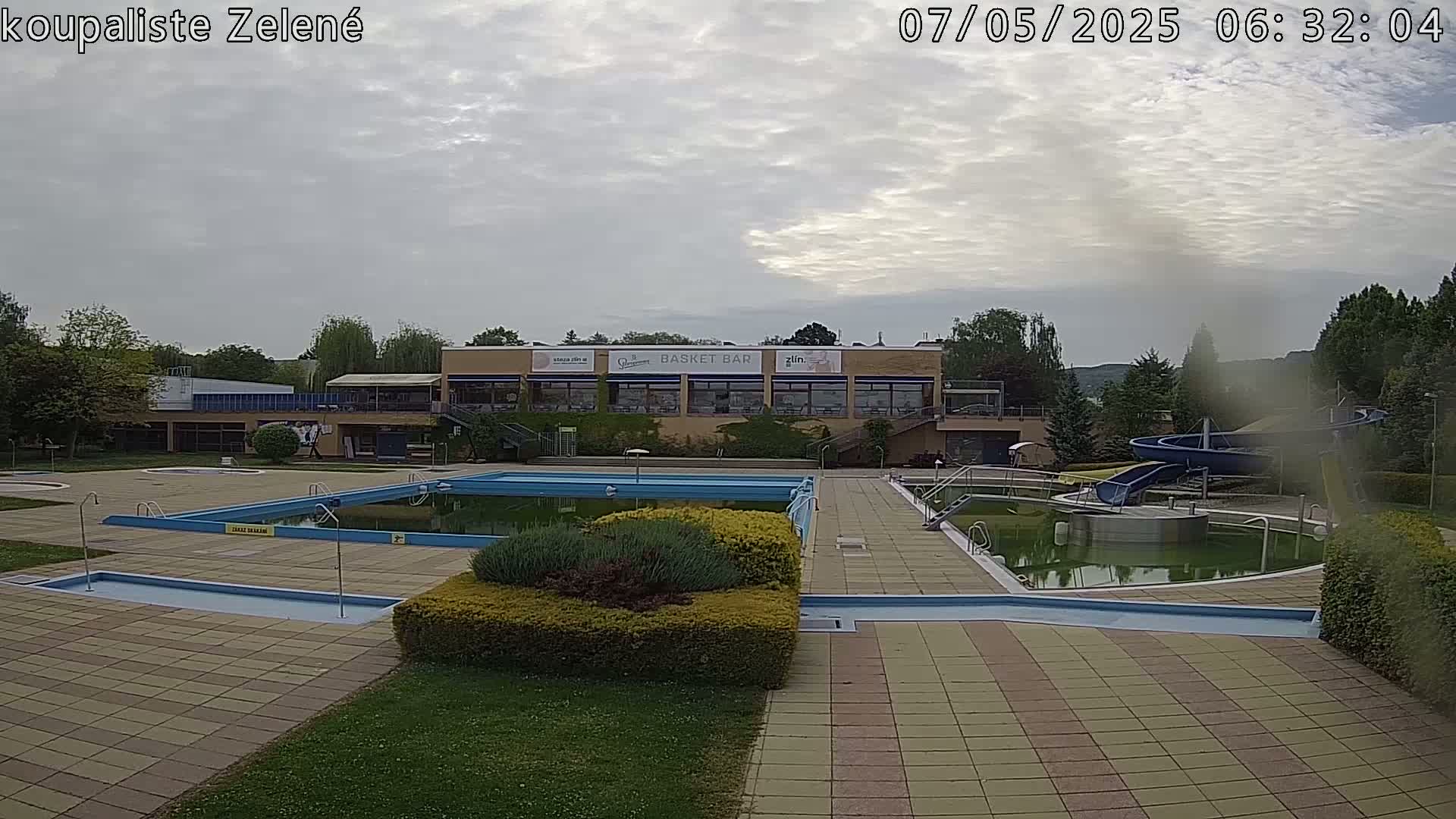 An outdoor swimming pool complex with multiple pools, a water slide, and a building with a restaurant under an overcast sky.