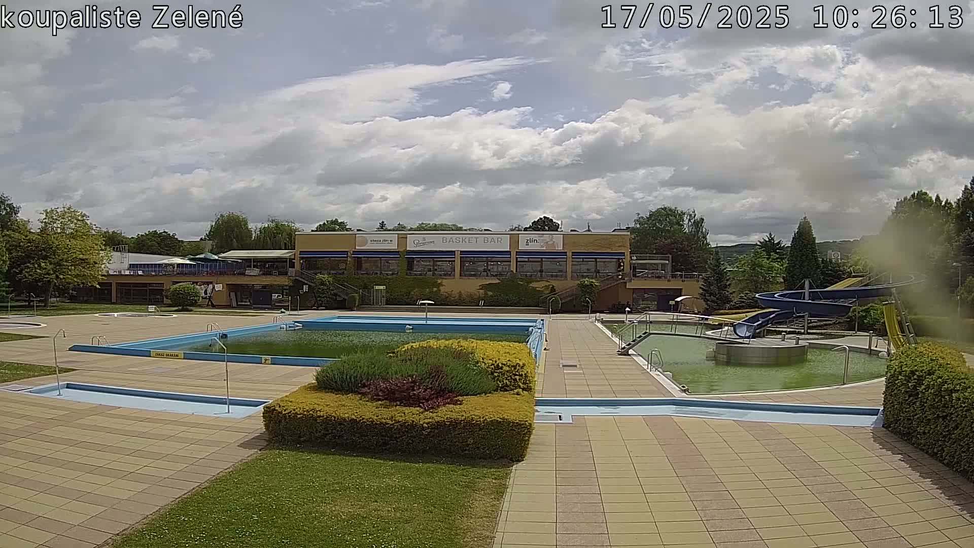 An outdoor swimming pool complex with multiple pools, a water slide, and a building, under a partly cloudy sky.