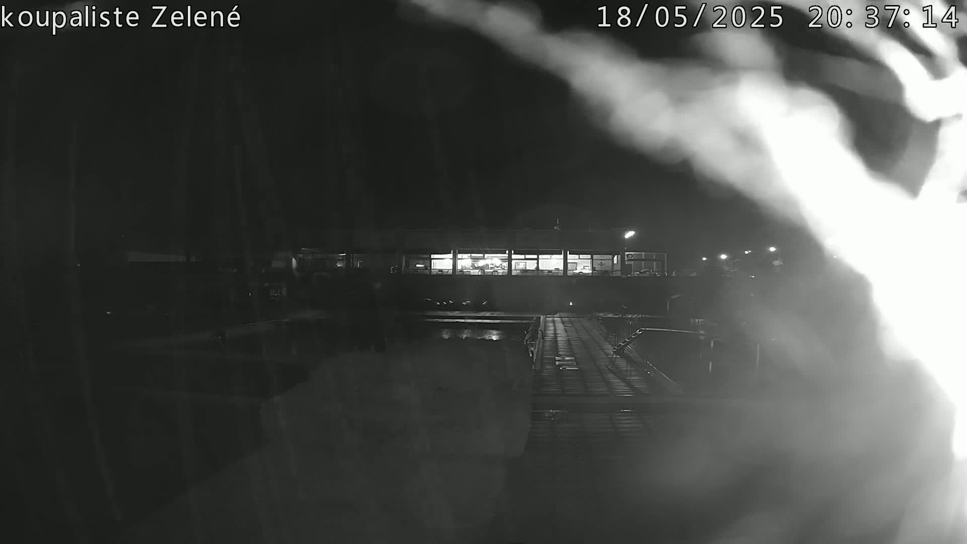 A nighttime, blurry black-and-white image shows a pool area with a building in the background, possibly steam or fog obscuring the view.