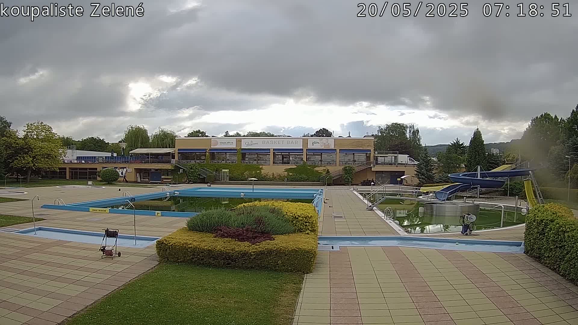 An outdoor swimming pool complex with a building, water slides, and landscaping under a partly cloudy sky.