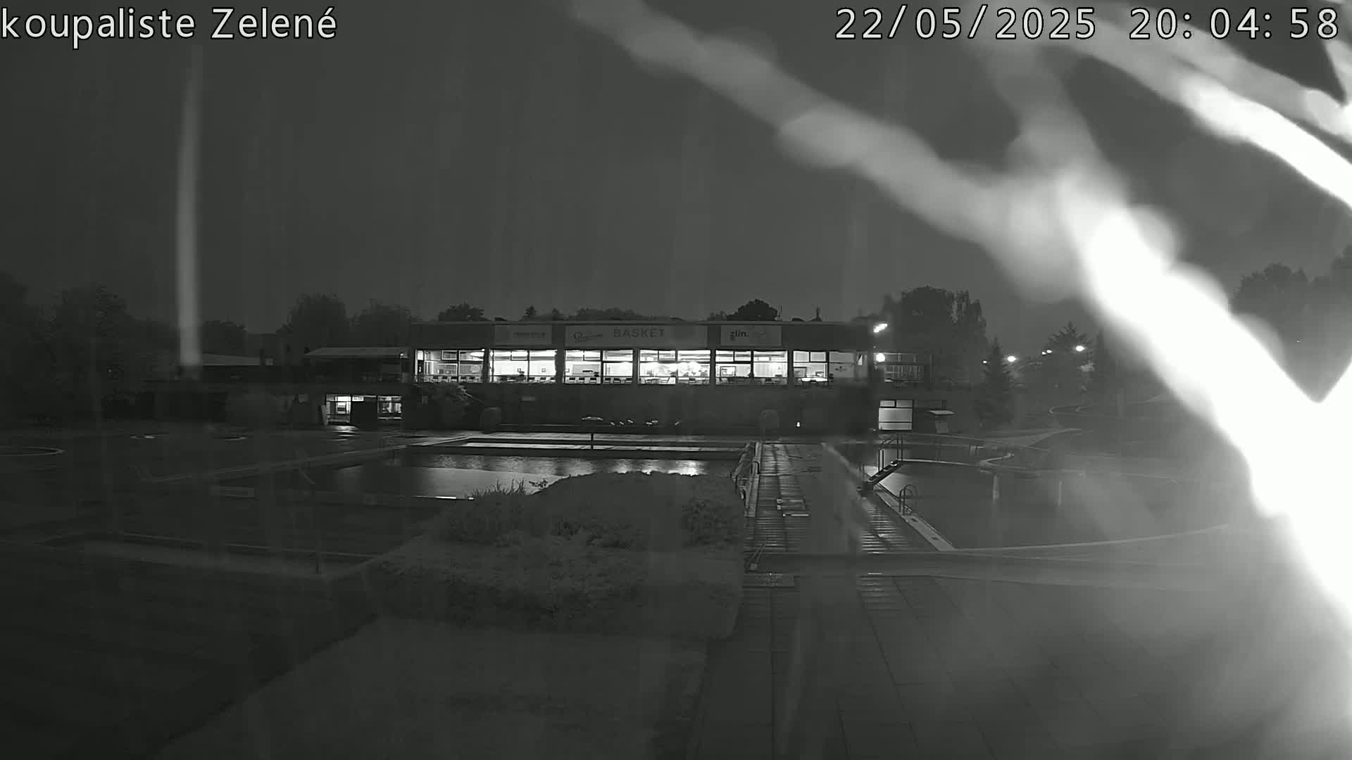 A nighttime, rainy view of an outdoor swimming pool complex with a building containing illuminated windows.