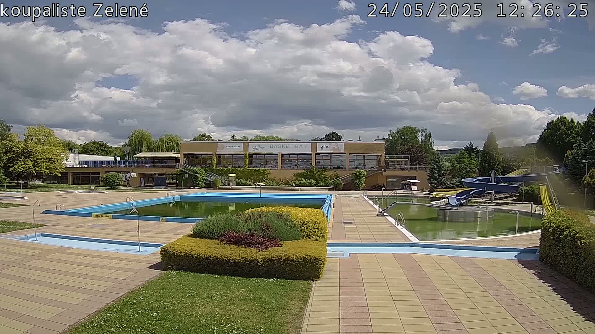 A partly cloudy outdoor scene shows an empty swimming pool complex with water slides and a building in the background.