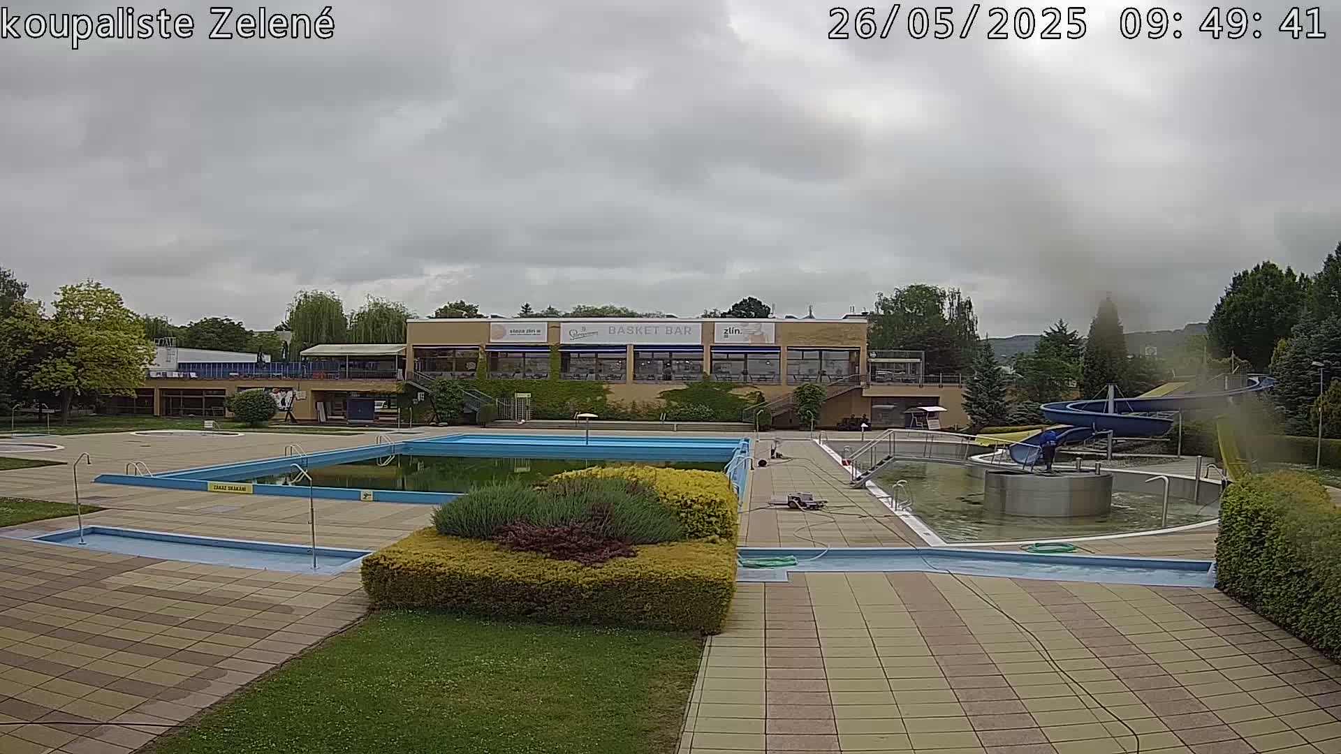 An outdoor swimming pool complex with a water slide is shown under an overcast sky.