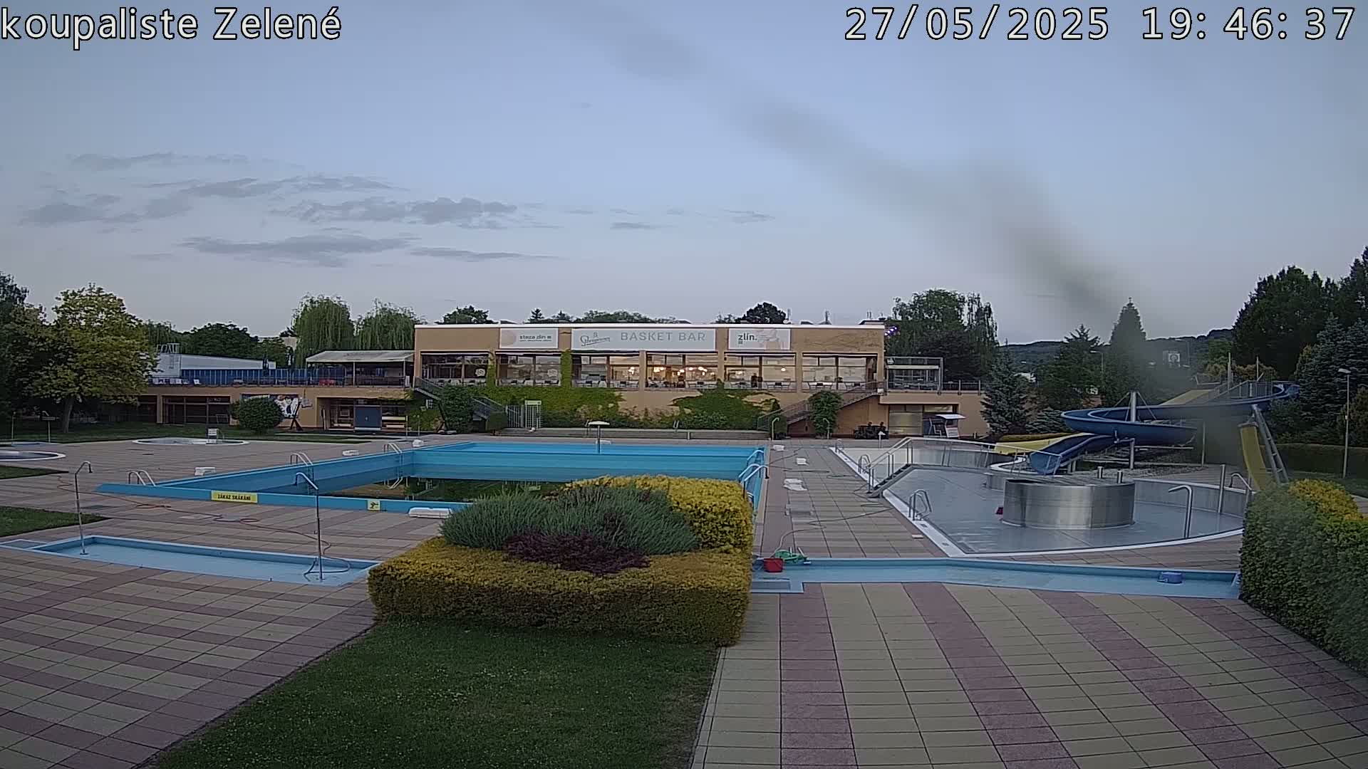 An outdoor pool complex with a large pool, a smaller pool, a water slide, and a building with a restaurant under an overcast sky.