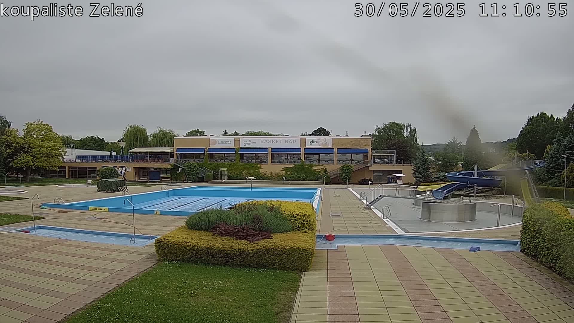 An outdoor swimming pool complex with a large rectangular pool, a smaller pool, and a water slide, under an overcast sky.