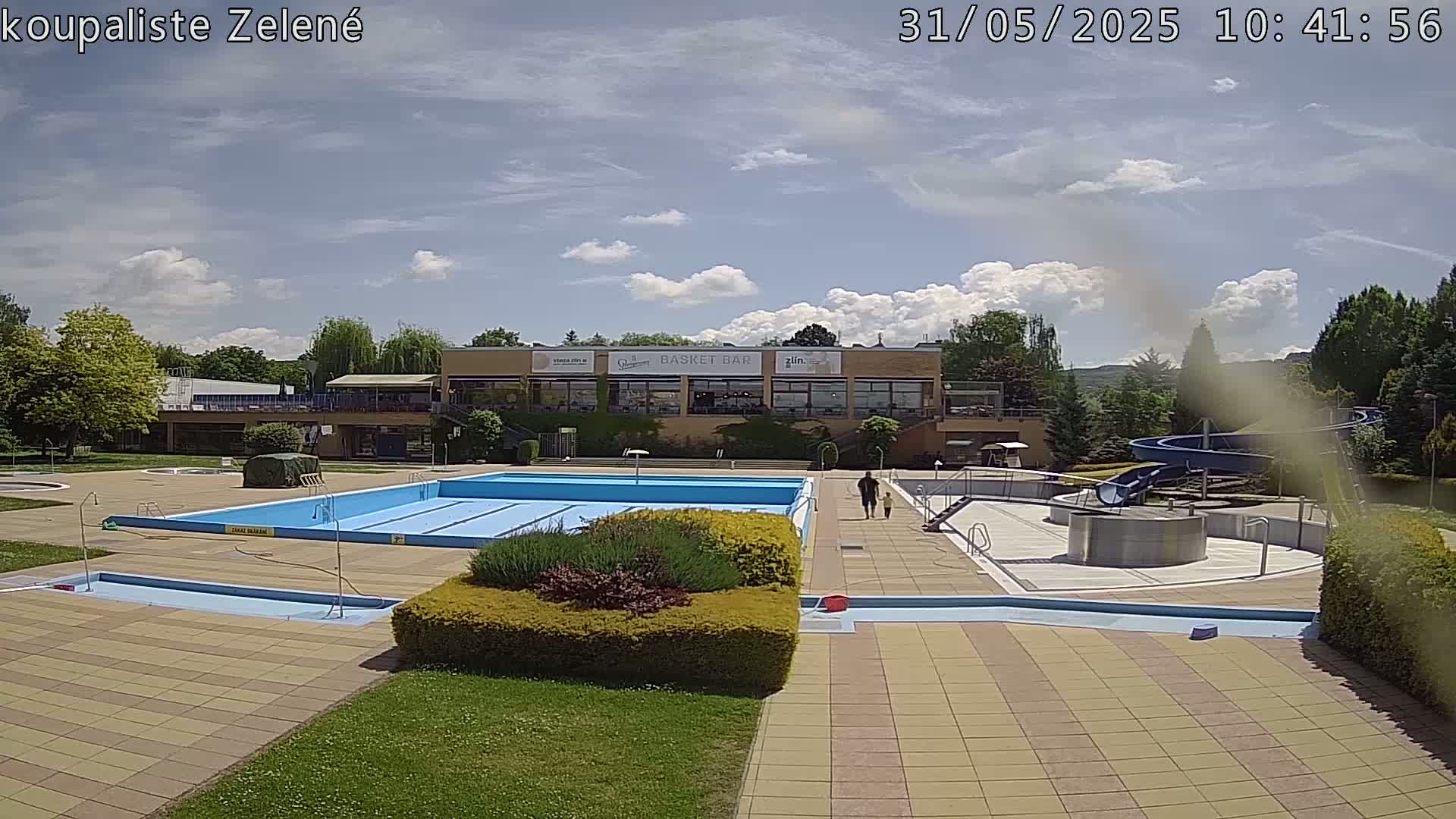 A sunny outdoor swimming pool complex with a large rectangular pool, a smaller children's pool, a water slide, and a building with a restaurant.