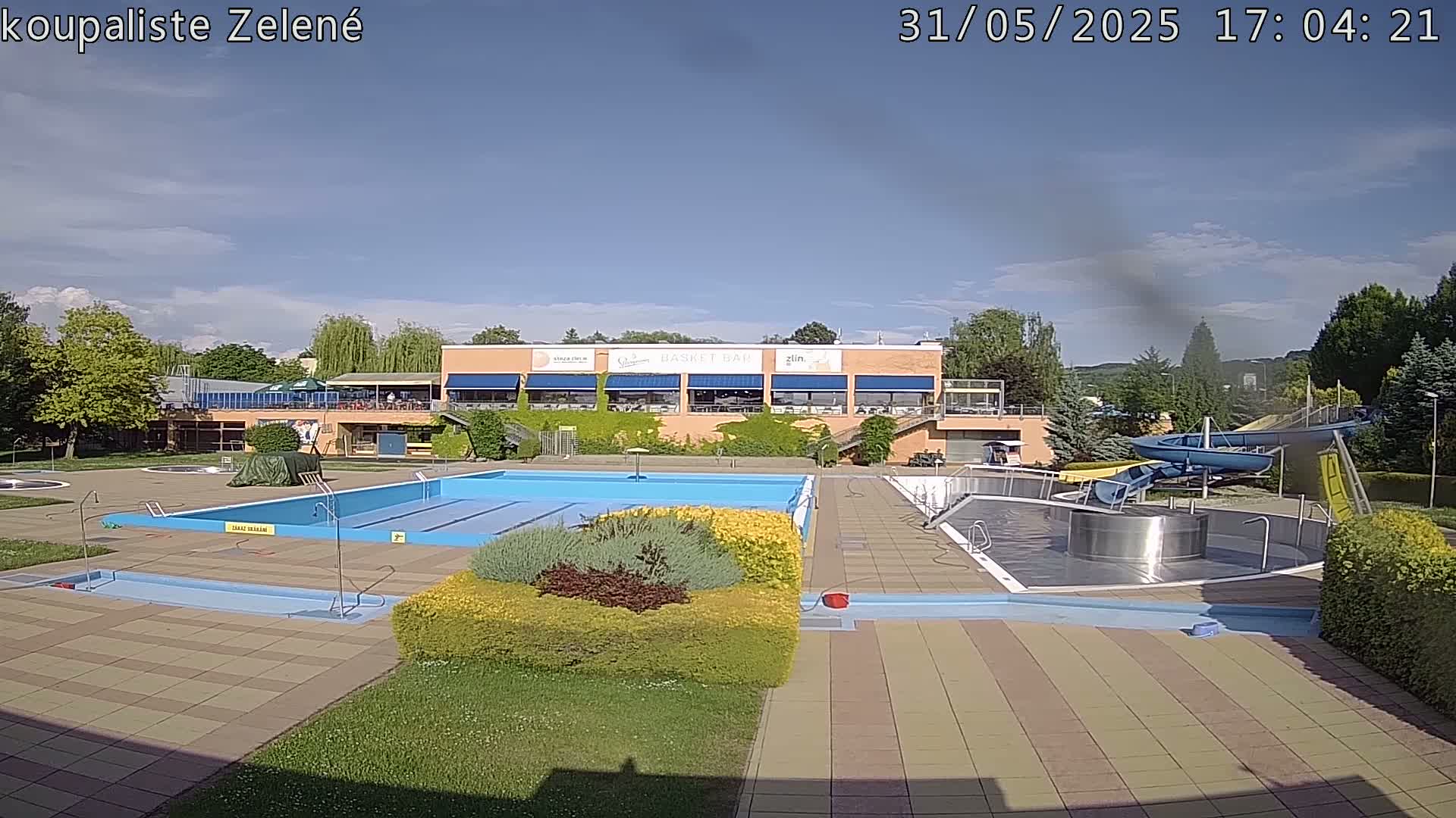 An outdoor swimming pool complex with a large main pool, smaller pools, water slides, and a building with a restaurant under a partly cloudy sky.