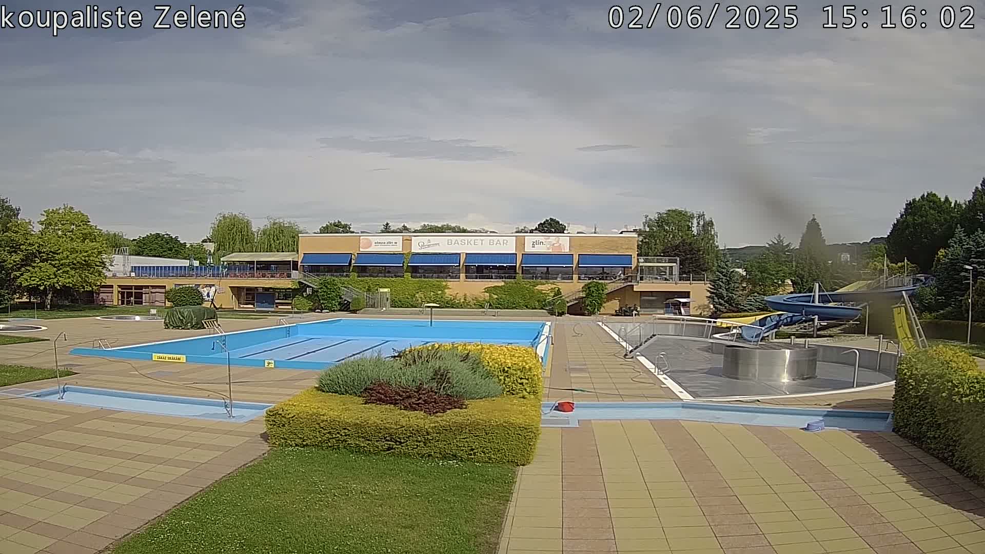 An outdoor swimming pool complex with a large swimming pool, a smaller pool, a water slide, and a building with a snack bar, under partly cloudy skies.