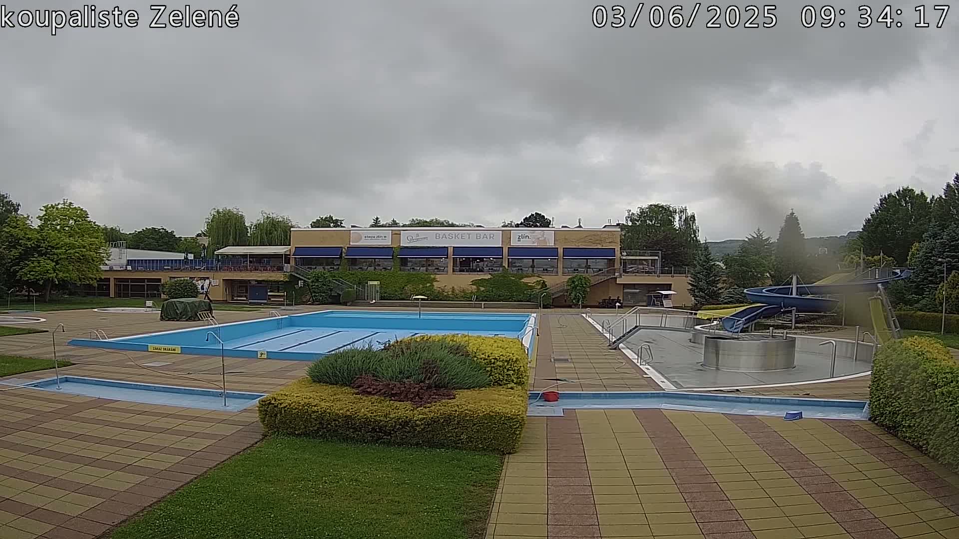 An outdoor swimming pool complex with a large main pool, smaller pools, and water slides sits under an overcast sky.