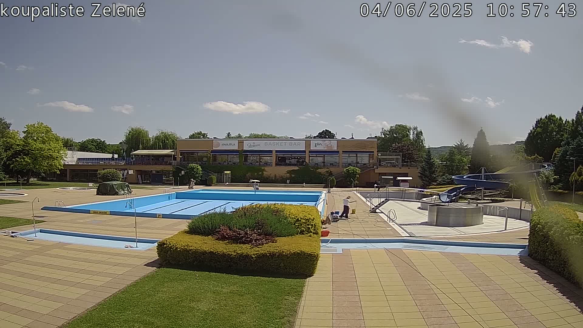 A sunny outdoor pool complex with a large swimming pool, a smaller wading pool, a water slide, and a building with a restaurant.