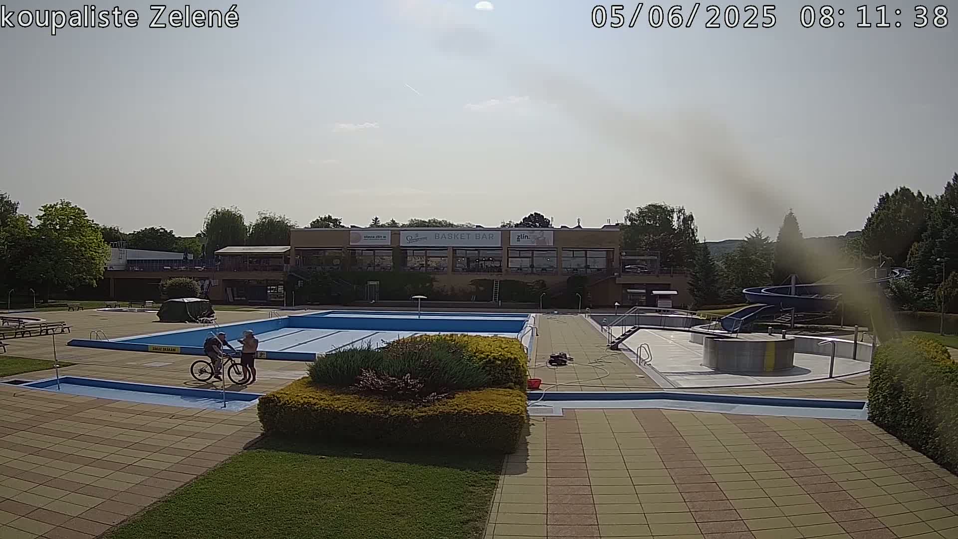 An outdoor swimming pool complex on a partly sunny day, with two people standing near bicycles next to the main pool, and a water slide visible in the background.