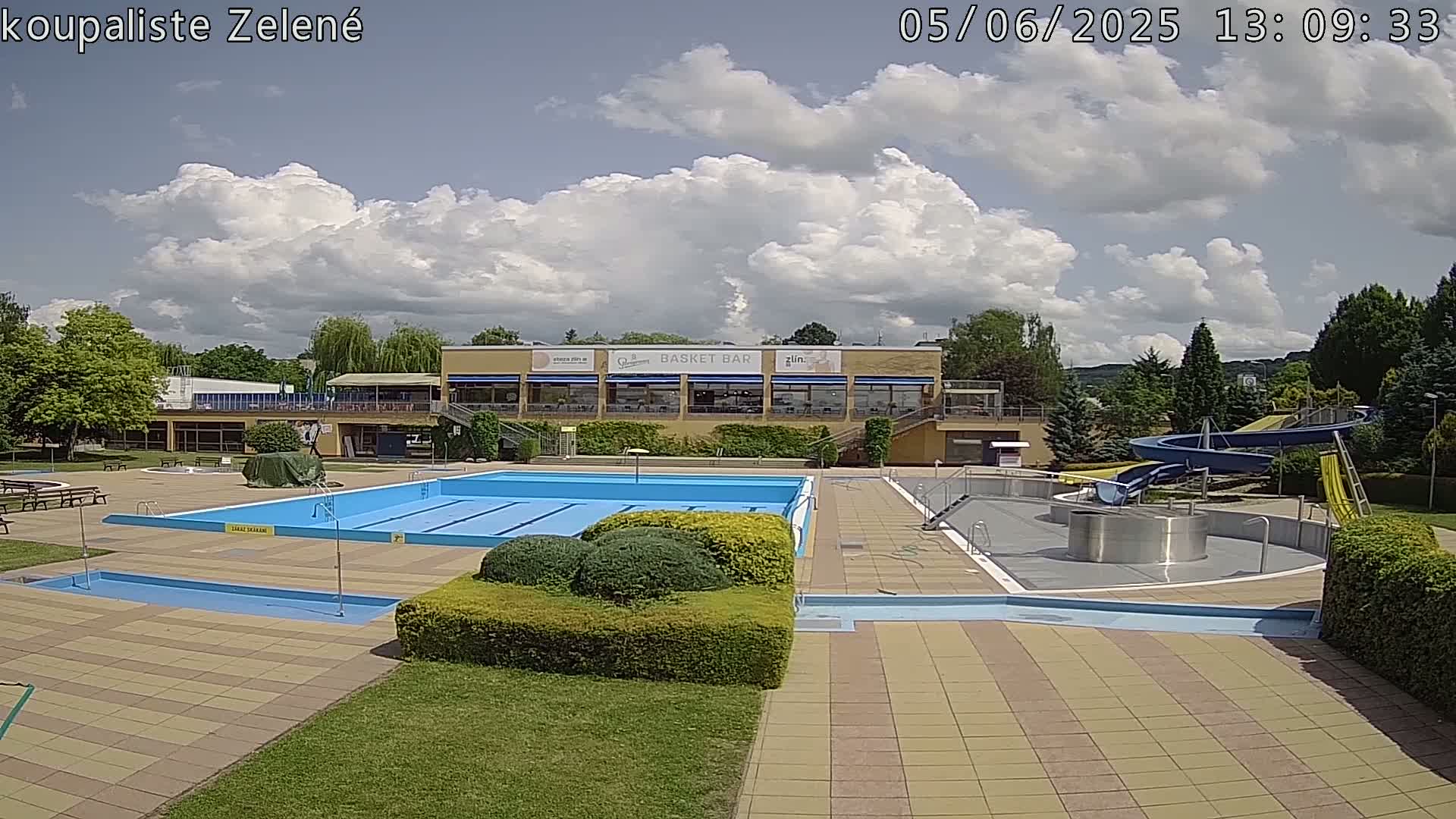 An outdoor swimming pool complex with a large main pool, smaller children's pool, water slides, and a building with a restaurant under a partly cloudy sky.