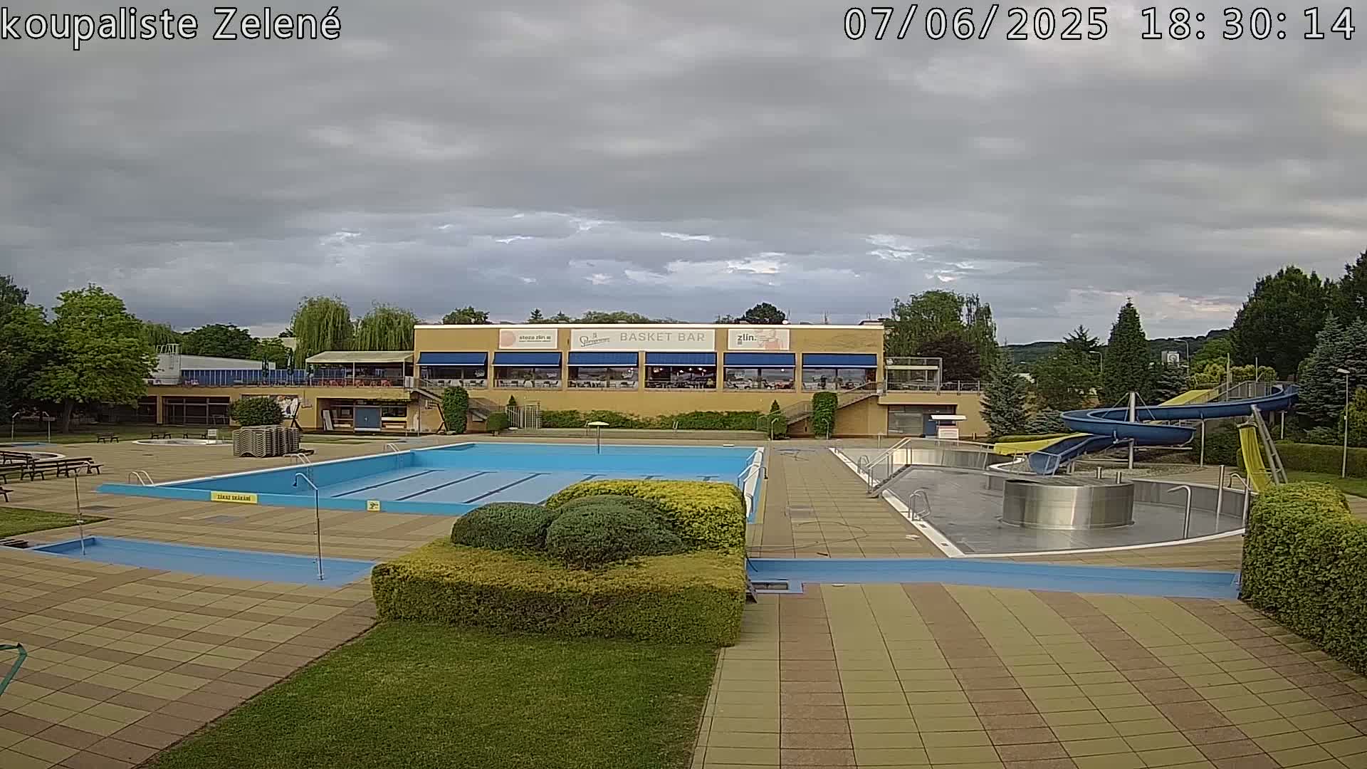 An outdoor swimming pool complex with a large pool, water slides, and a building with a restaurant under a cloudy sky.
