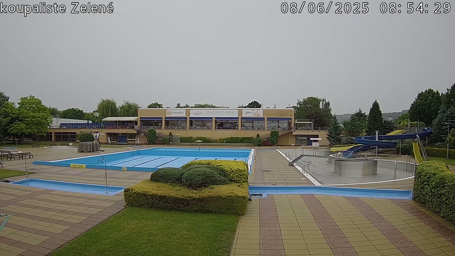 An outdoor swimming pool complex with a large swimming pool, a smaller wading pool, water slides, and a building with a restaurant is shown under an overcast sky.