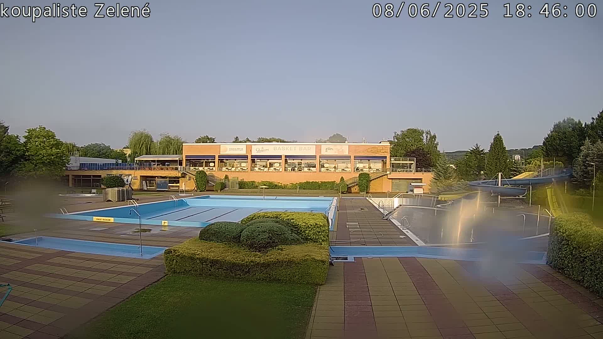 An outdoor swimming pool complex with a large rectangular pool, a smaller wading pool, a water slide, and a building with a restaurant.