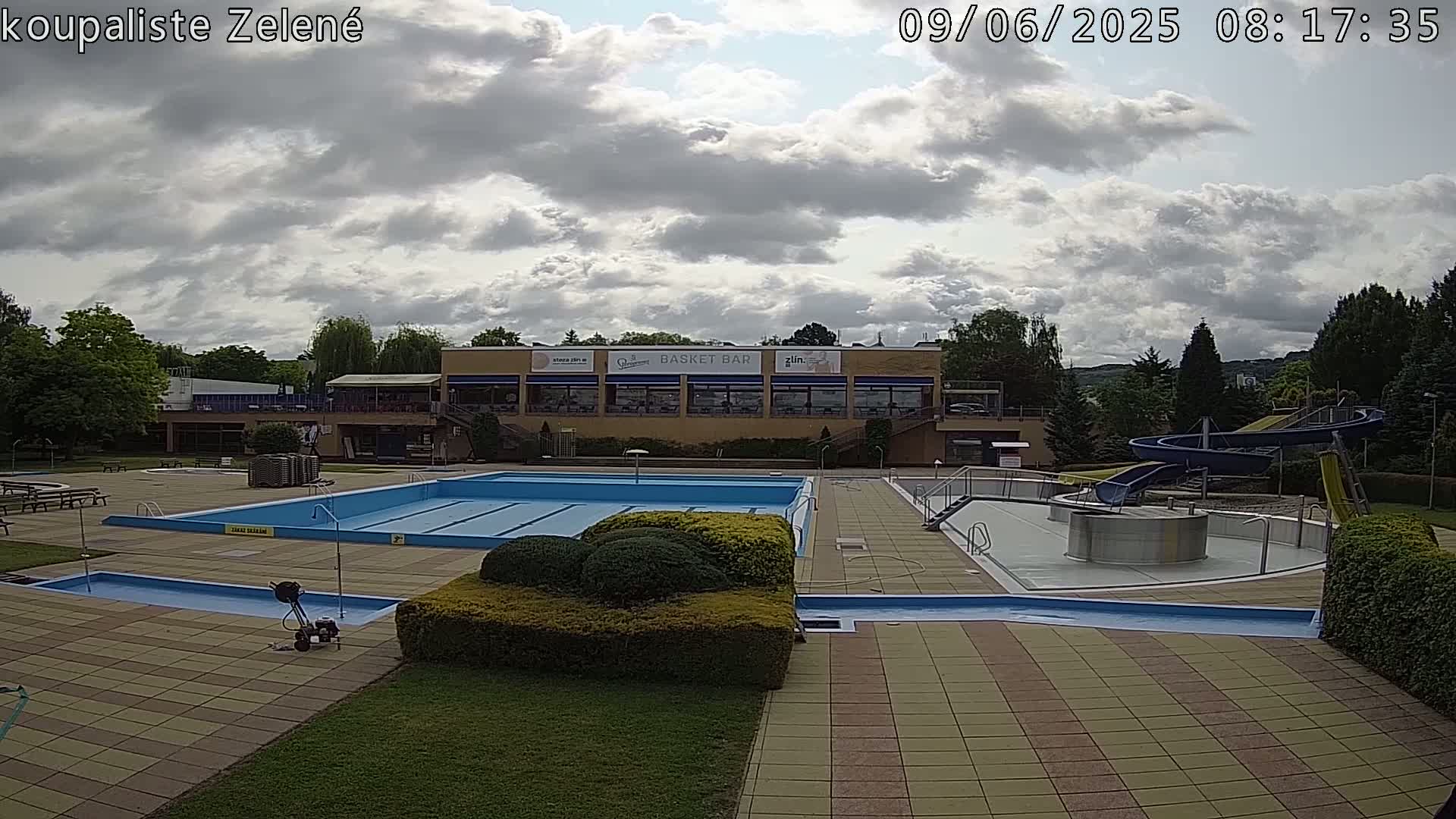 An outdoor swimming pool complex with a large main pool, smaller pools, water slides, and a building with a restaurant under a partly cloudy sky.
