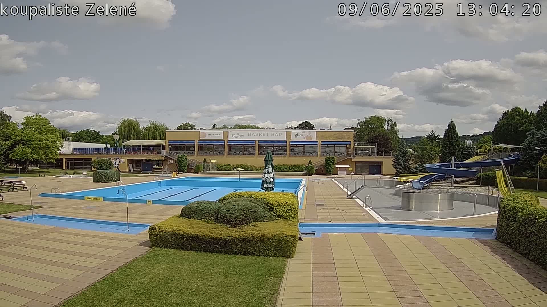 A sunny outdoor swimming complex with a large pool, a smaller pool, water slides, and a building with a restaurant.