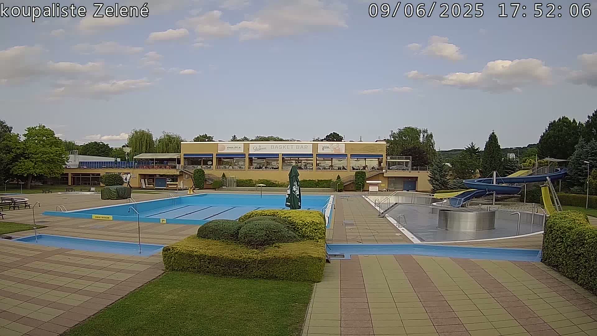 An outdoor pool complex with a large swimming pool, water slides, and a building with a restaurant under a partly cloudy sky.