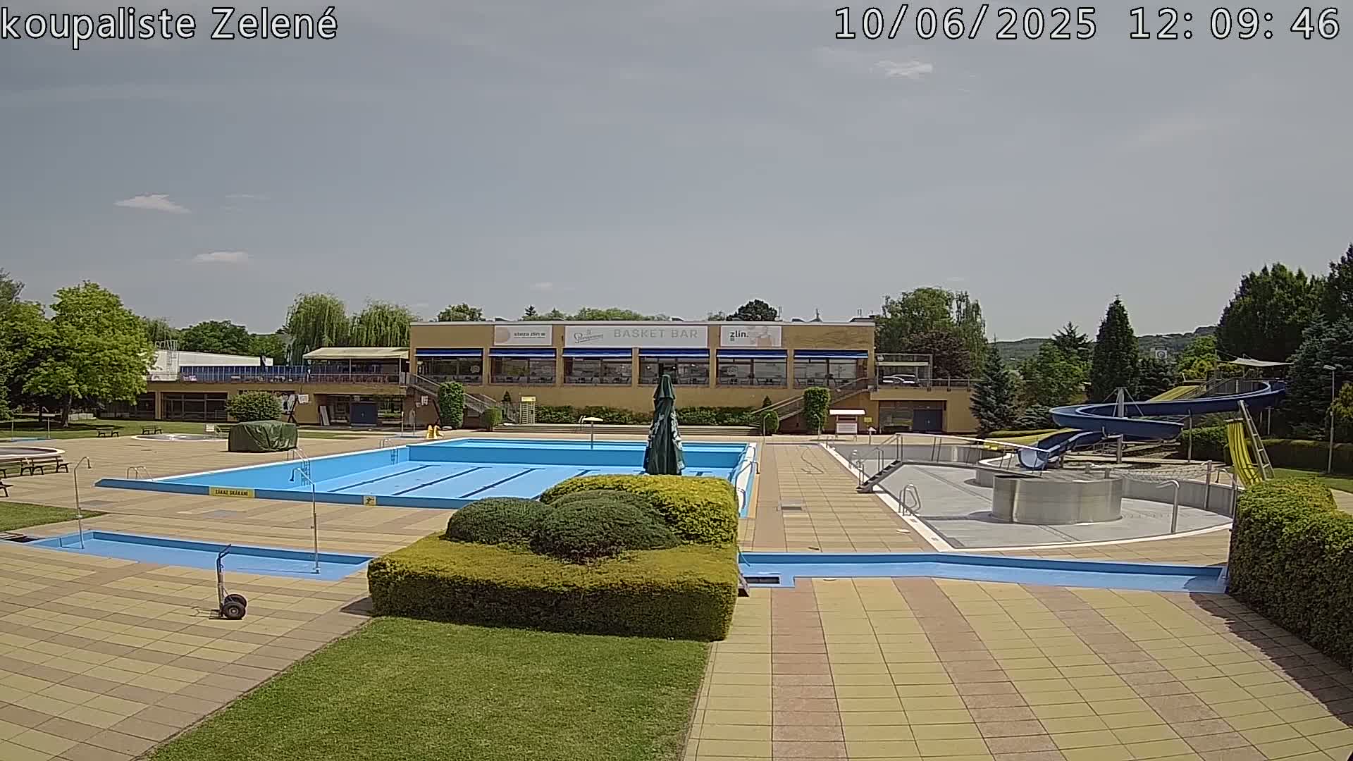 An outdoor swimming pool complex with a large rectangular pool, smaller pools, and a water slide, under a mostly sunny sky.