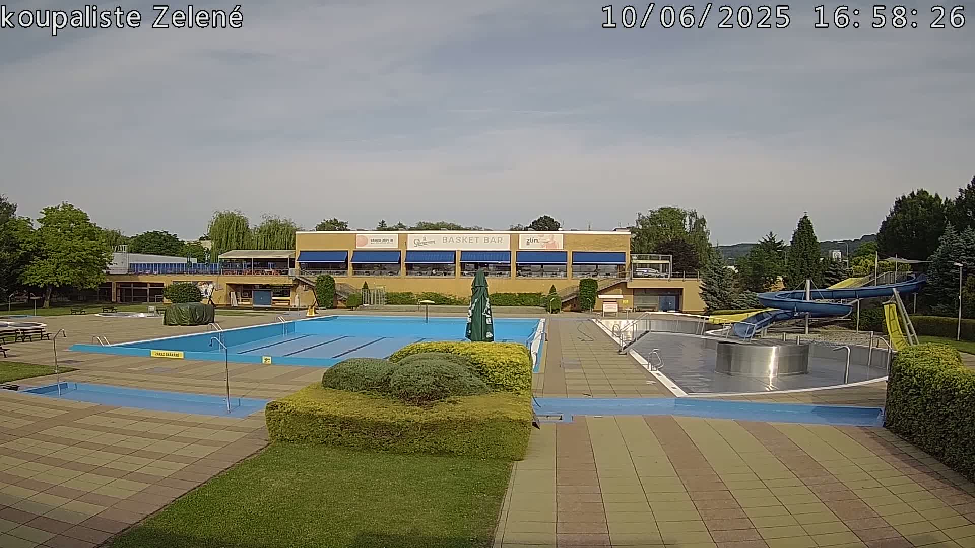 An outdoor swimming pool complex with a large main pool, smaller pools, water slides, and a building with a restaurant on a partly cloudy day.