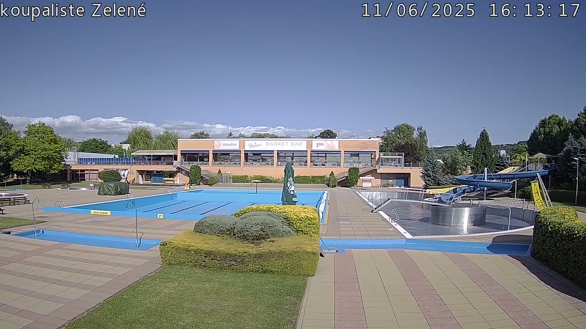 An outdoor swimming pool complex with a large pool, water slides, and a building with a restaurant under mostly sunny skies.