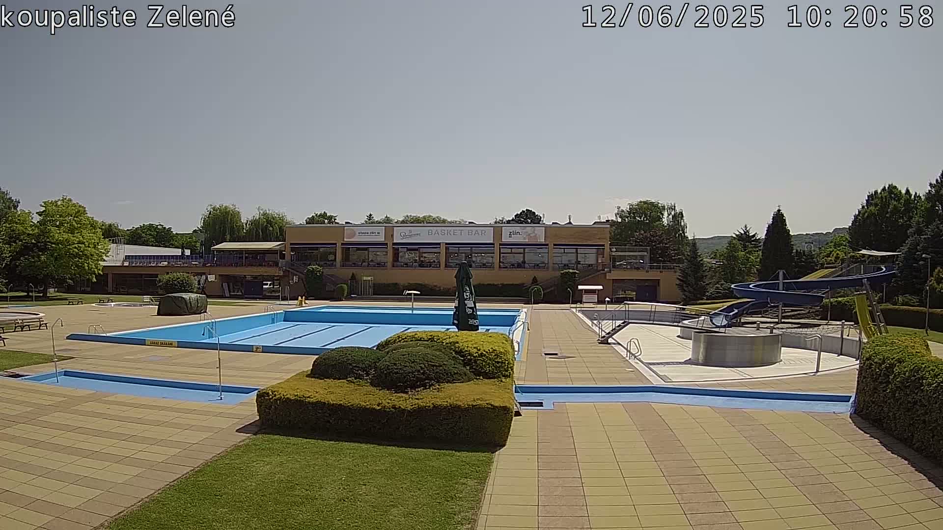 An outdoor pool complex with a large swimming pool, a smaller wading pool, a water slide, and a building with a restaurant in the background under a sunny sky.